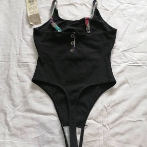 May include: Black bodysuit with a scoop neck and thin straps. The bodysuit has a button closure at the front and a thong back.