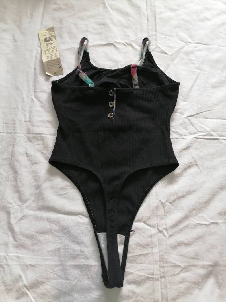 May include: Black bodysuit with a scoop neck and thin straps. The bodysuit has a button closure at the front and a thong back.