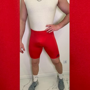 Può includere: Un top bianco senza maniche a girocollo abbinato a pantaloncini sportivi a compressione rosso acceso con un piccolo logo Nike nero sulla gamba sinistra. L'abbigliamento è completato da calzini bianchi e scarpe da ginnastica grigie con catena.
