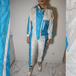 Puede incluir: Un traje de esquí blanco y azul con cierre de cremallera y un diseño de color contrastante. El traje tiene un cuello alto y mangas largas.