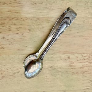 Könnte beinhalten: Vintage, versilberte Zuckerklemme mit löffelförmigen Enden. Die Zange hat ein verziertes Design an den Griffen und liegt auf einer hellbraunen Holzoberfläche. Die Zange ist leicht geöffnet.