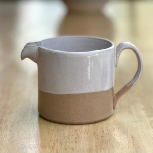 May include: A two-tone ceramic jug with a handle and a spout. The top half is a light cream colour, and the bottom half is a tan colour. The jug is sitting on a wooden surface.