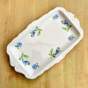 May include: A rectangular white ceramic tray with scalloped edges and a delicate floral pattern featuring small blue flowers with green stems and leaves scattered across the surface. The tray has a glossy finish and a slightly raised, textured border.