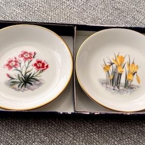 May include: Two small, white porcelain dishes with gold rims, each featuring a floral design. One dish has red flowers, the other yellow. They are presented in a dark blue box, ideal for serving small portions.