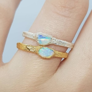 Puede incluir: Dos anillos con piedras preciosas de ópalo. El anillo superior es de plata con un acabado texturizado. El anillo inferior es de oro con un acabado texturizado.