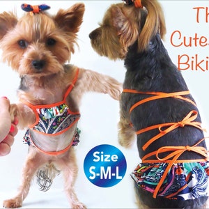 May include: A small dog wearing a colorful bikini with orange straps and a paisley print. The dog is standing on a white background. The text "The Cutest Bikini" and "Size S-M-L" are visible in the image.