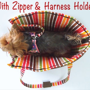 May include: A striped fabric dog carrier with a zipper and a harness holder. A small dog is inside the carrier.
