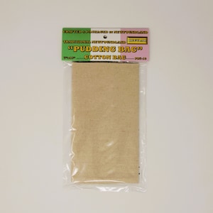 Peut inclure: Un sac à pudding en coton beige, emballé dans du plastique transparent. L'étiquette en haut indique "PUDDING BAG" et "COTTON BAG". L'étiquette indique également "Crafted & Packaged in Newfoundland". Le sac mesure 18 x 25 cm.