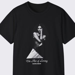 May include: Black t-shirt featuring a black and white image of a person with long hair. The text "The Art of Loving" and the name "Olivia Dean" are printed in white script below the image.