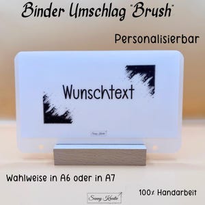 Könnte beinhalten: Eine weiße Ordnerhülle mit dem Text "Wunschtext" und schwarzen Pinselstrich-Akzenten. Die Hülle wird auf einem Holzsockel präsentiert. Der Text "Binder Umschlag Brush", "Personalisierbar", "Wahlweise in A6 oder in A7" und "100% Handarbeit" sind ebenfalls sichtbar.