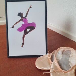 May include: A framed print of a black ballerina in a pink tutu, with a pair of ballet slippers on a wooden surface.