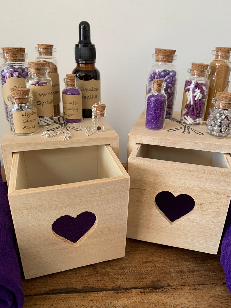 Puede incluir: Dos cajas de madera con recortes de coraz&oacute;n morado, cada una contiene peque&ntilde;os frascos de vidrio llenos de varios espolvoreados morados y plateados, y una botella de extracto de vainilla. Los frascos est&aacute;n etiquetados con nombres como "Elysian Silver Stars", "Werphan Sprinkles" y "Kalimba Baking Sugar".