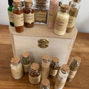 Puede incluir: Una caja de madera con tapa abatible, que contiene pequeños frascos de vidrio llenos de diversas hierbas, polvos y otros ingredientes. Los frascos están etiquetados con nombres escritos a mano, como "Witch's Cackle", "Feather of Flamingo" y "White Hair of Hare".