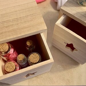 Puede incluir: Dos cajas de madera con cajones, cada una con un recorte en forma de estrella. El caj&oacute;n superior de la caja de la izquierda est&aacute; abierto, revelando cinco peque&ntilde;as botellas de vidrio con tapas de corcho llenas de purpurina de colores.