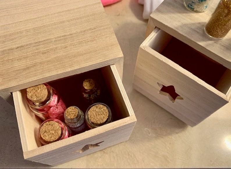 Puede incluir: Dos cajas de madera con cajones, cada una con un recorte en forma de estrella. El caj&oacute;n superior de la caja de la izquierda est&aacute; abierto, revelando cinco peque&ntilde;as botellas de vidrio con tapas de corcho llenas de purpurina de colores.