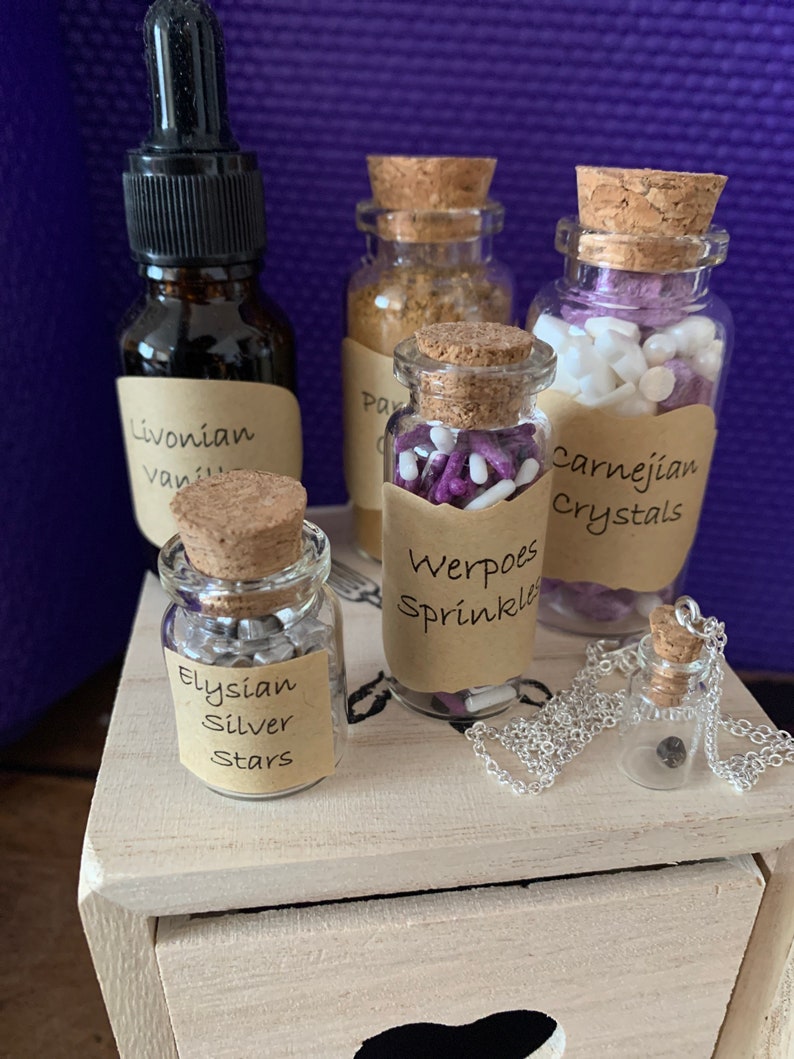 Puede incluir: Una caja de madera con cinco peque&ntilde;os frascos de vidrio que contienen varios art&iacute;culos. Los frascos tienen tapones de corcho y etiquetas con nombres como "Livonian Vanilla", "Werpoes Sprinkles", "Elysian Silver Stars" y "Carnelian Crystals".