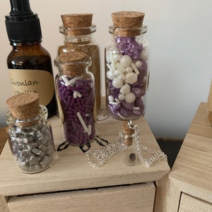 Puede incluir: Cinco peque&ntilde;os frascos de vidrio con tapas de corcho llenos de diferentes tipos de espolvorear y un collar de plata con un peque&ntilde;o colgante de frasco de vidrio. Los frascos est&aacute;n dispuestos sobre una superficie de madera con un recorte en forma de coraz&oacute;n.