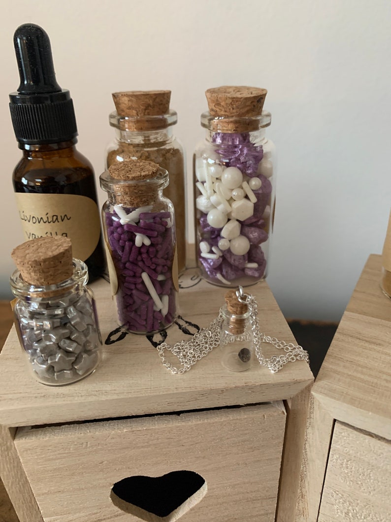 Puede incluir: Cinco peque&ntilde;os frascos de vidrio con tapas de corcho llenos de diferentes tipos de espolvorear y un collar de plata con un peque&ntilde;o colgante de frasco de vidrio. Los frascos est&aacute;n dispuestos sobre una superficie de madera con un recorte en forma de coraz&oacute;n.