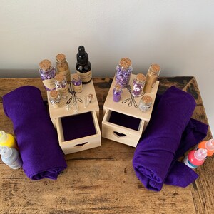 Puede incluir: Dos cajas de madera con cajones, cada una con peque&ntilde;as botellas de vidrio llenas de purpurina morada y dorada. Las cajas est&aacute;n sobre una mesa de madera con dos rollos de tela morada y dos botellas de pintura.