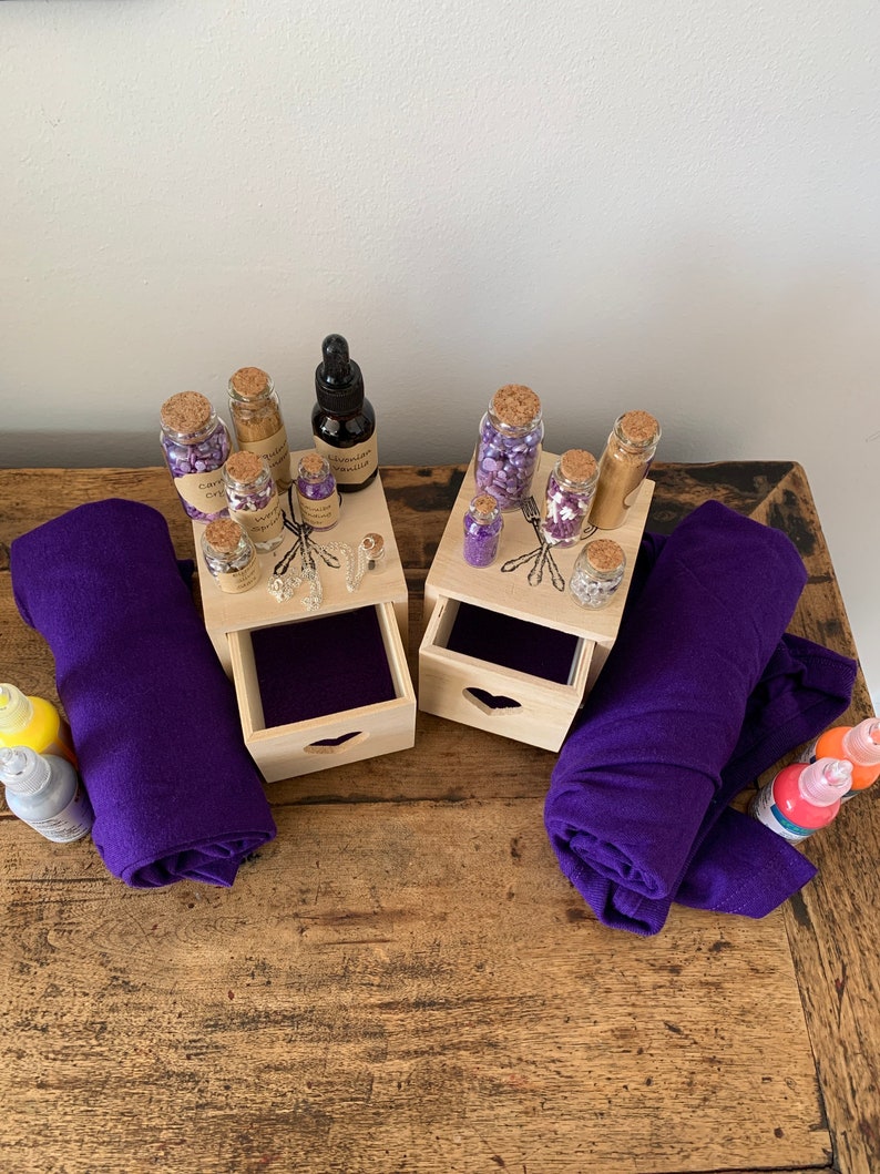 Puede incluir: Dos cajas de madera con cajones, cada una con peque&ntilde;as botellas de vidrio llenas de purpurina morada y dorada. Las cajas est&aacute;n sobre una mesa de madera con dos rollos de tela morada y dos botellas de pintura.