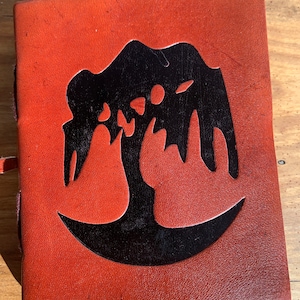 May include: A brown leather journal with a black tree design embossed on the front cover.