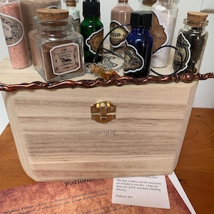 Könnte beinhalten: Eine Holzkiste mit einem Messingverschluss, beschriftet mit "Property of" und gefüllt mit kleinen Glasflaschen, die verschiedene farbige Pulver und Flüssigkeiten enthalten. Die Flaschen sind mit Namen wie "Unicorn Blood", "Tears" und "Polyjuice Potion" beschriftet. Ein Zettel rechts lautet "This class is remote and the instructions are included in your box. I hope you have a lot of fun, and make something delicious." Professor Jam