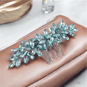 May include: A silver hair comb with a decorative design of light blue iridescent crystals. The crystals are arranged in a floral pattern.