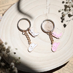 Könnte beinhalten: Zwei Schlüsselanhänger mit silbernen Ringen, goldenen Ketten und Charms im Cowboy-Stil. Ein Schlüsselanhänger hat weiße Charms von einem Cowboy-Stiefel und -Hut. Der andere hat rosa Charms. Auf einer Holzoberfläche.