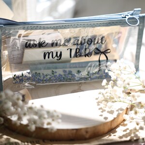 Può includere: Astuccio trasparente con cerniera blu e testo nero "Ask me about my TBR". Contiene vari oggetti e un motivo floreale blu. Fiori bianchi e una casa decorativa sullo sfondo, su una superficie di legno.