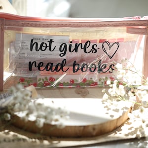 Könnte beinhalten: Transparente Federtasche mit rosa Einfassung, mit dem Text "hot girls read books" und einem Herz. Die Tasche enthält Schreibwaren und dekorative Elemente, umgeben von Blumen.