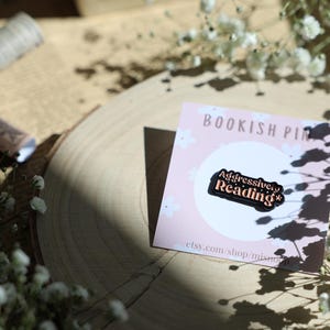 Könnte beinhalten: Eine schwarze Emaille-Anstecknadel mit dem Text "Aggressively Reading*" in rosa und weiß. Die Anstecknadel befindet sich auf einer rosa Karte mit dem Text "Bookish Pin" und einer Website-Adresse.