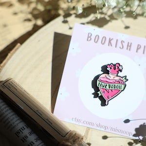 Könnte beinhalten: Eine rosa und grüne Emaille-Anstecknadel in Form einer herzförmigen Flasche mit einem Etikett, auf dem "Love Potion" steht. Die Anstecknadel befindet sich auf einer rosa Karte mit dem Text "Bookish Pin" und einer Website-Adresse.