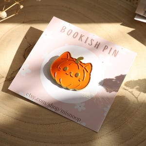Può includere: Spilla in smalto a forma di zucca arancione con un volto sorridente e un gambo verde, esposta su una carta rosa con la scritta "BOOKISH PIN". La spilla è appoggiata su una superficie di legno, con l'URL del negozio Etsy visibile.