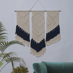 May include: A wall hanging made of macrame with a geometric pattern. The macrame is white with black fringe. The wall hanging is made of three panels.