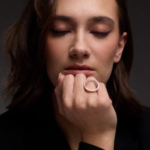 May include: A silver ring with a circular design. The ring is worn on a finger. The image is a close-up shot of the ring and the person's hand. The background is a dark gray.