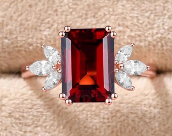 Op de afbeelding: Een roségouden ring met een grote, rechthoekige, smaragdgeslepen dieprode granaat in het midden, geflankeerd aan weerszijden door drie kleine, marquise-geslepen helderwitte stenen die in een waaiervorm op de dunne metalen ringband zijn geplaatst.