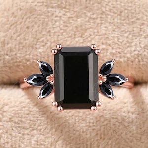 K&ouml;nnte beinhalten: Ein Ring aus Ros&eacute;gold mit einem gro&szlig;en, schwarz gef&auml;rbten Stein im Smaragdschliff, der auf jeder Seite von drei schwarzen Steinen im Marquiseschliff flankiert wird. Der Ring hat eine Krappenfassung, eine polierte Metallschiene und dunkle Edelsteine.