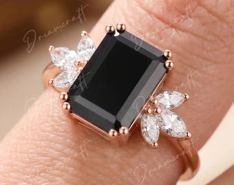 May include: A rose gold ring featuring a large rectangular black center stone flanked by two clusters of three marquise-cut clear stones on each side. The band is thin and polished, holding the central black gemstone in a prong setting.