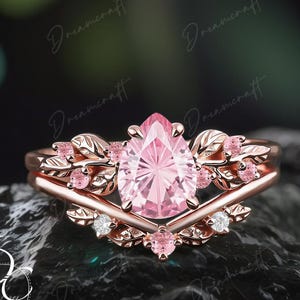 May include: A rose gold engagement ring set with a pear-shaped pink gemstone. The ring is adorned with leaf and small pink stone accents. The band is rose gold with a matching wedding band.