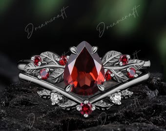May include: A black metal ring set featuring a pear-shaped red garnet center stone, accented with smaller round red garnets and clear stones. The band is designed with intricate leaf motifs and a V-shaped contour, creating a layered appearance.