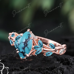 May include: Rose gold ring featuring a large, teardrop-shaped turquoise stone with black veining. The ring is adorned with smaller turquoise accents and clear, marquise-cut stones, set in a floral design. The ring is displayed on a dark, textured surface.