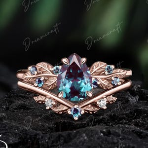 May include: A rose gold ring set featuring a pear-cut color-changing center stone, accented by smaller round stones and leaf-shaped metal details. The band has a V-shaped contour, and the design incorporates intricate botanical-inspired engravings.