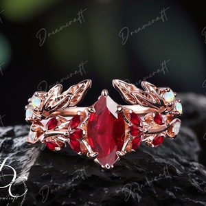 May include: A rose gold ring with a large red gemstone surrounded by smaller red gemstones and white opals. The ring features a delicate leaf design.
