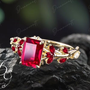 Peut inclure: Une bague en or ornée d'une grande pierre rouge rectangulaire au centre, entourée de petites pierres rouges et de délicats motifs de feuilles d'or. La bague est posée sur une surface sombre et texturée.
