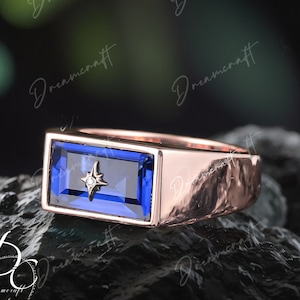 Può includere: Un anello in oro rosa con una pietra preziosa blu rettangolare incastonata in una montatura a becco di lumaca. Un piccolo diamante è incastonato al centro della pietra preziosa.