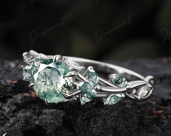 May include: A silver-toned ring featuring a round, moss-green gemstone with dark inclusions at the center. The band is designed with leaf-shaped accents and smaller green stones along the sides, creating a botanical-inspired metal setting.
