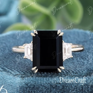 Könnte beinhalten: Ein goldener Ring mit einem großen, rechteckigen schwarzen Edelstein und zwei kleineren, rechteckigen weißen Edelsteinen. Der Ring ist vor einem blauen Hintergrund platziert. Der Text "DreamCraft" ist sichtbar.