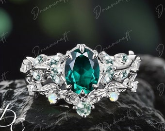 May include: A silver ring set with a large, oval-shaped emerald green gemstone surrounded by smaller, clear gemstones. The ring band is decorated with a delicate, leafy design.