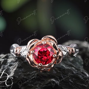May include: A rose-shaped ring with a red gemstone set in a rose gold band. The ring is surrounded by smaller white gemstones and has a silver band.