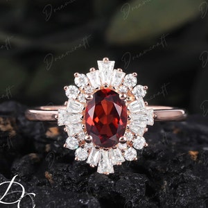 May include: A rose gold ring with a central oval-cut, deep red gemstone, encircled by a halo of smaller, clear baguette and round-cut stones. The ring's band is a delicate rose gold, and the design is intricate and eye-catching, suitable for special occasions.